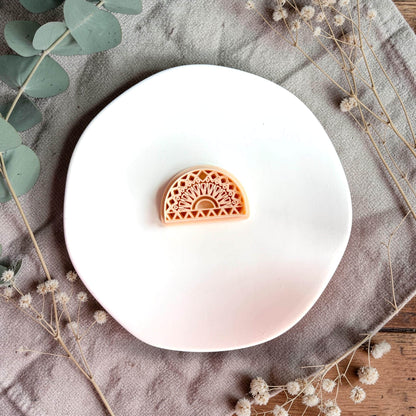 Small arch-shaped clay piece with intricate geometric sunburst pattern displayed on a white round plate with neutral fabric and greenery.