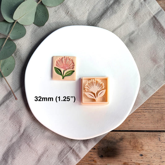 Square polymer clay cutter with pohutukawa flower design displayed on a white round plate with neutral fabric and greenery.