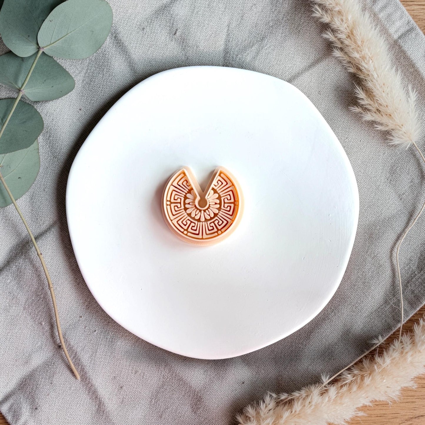 U-shaped polymer clay cutter with intricate Greek key and floral pattern displayed on a white round plate with neutral fabric.