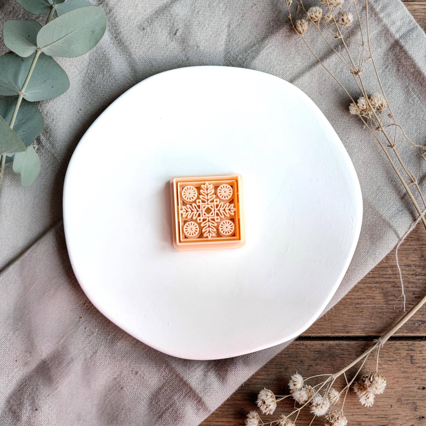 Square polymer clay cutter with intricate Nordic floral tile pattern displayed on a white round plate with neutral fabric and dried flowers.
