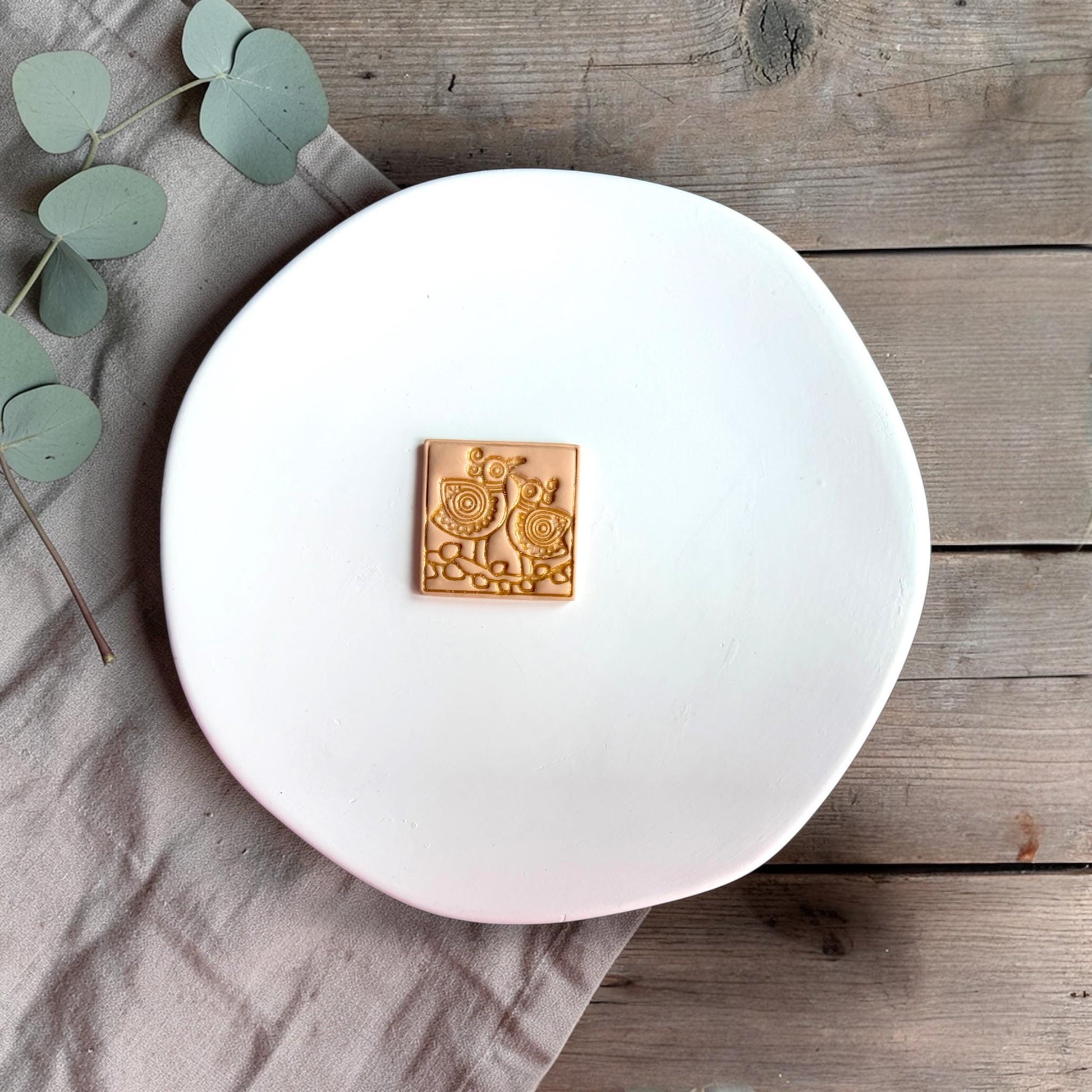 Small square clay piece with two stylized folk art birds embossed on the surface, placed on a white plate with neutral fabric.