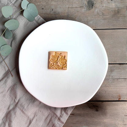 Small square clay piece with two stylized folk art birds embossed on the surface, placed on a white plate with neutral fabric.