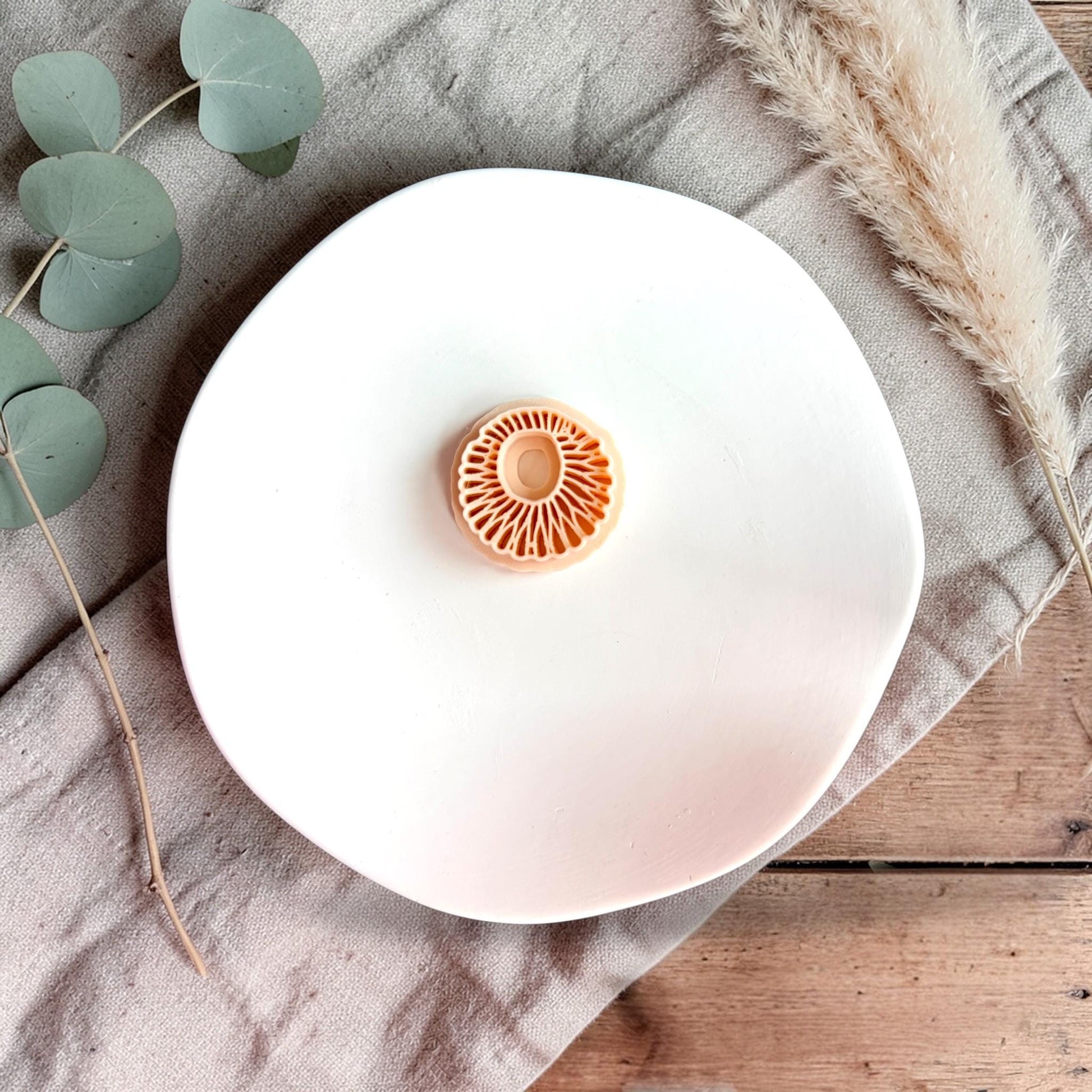 Small round polymer clay cutter with sunburst mandala pattern placed on a white plate with neutral fabric and dried grass.