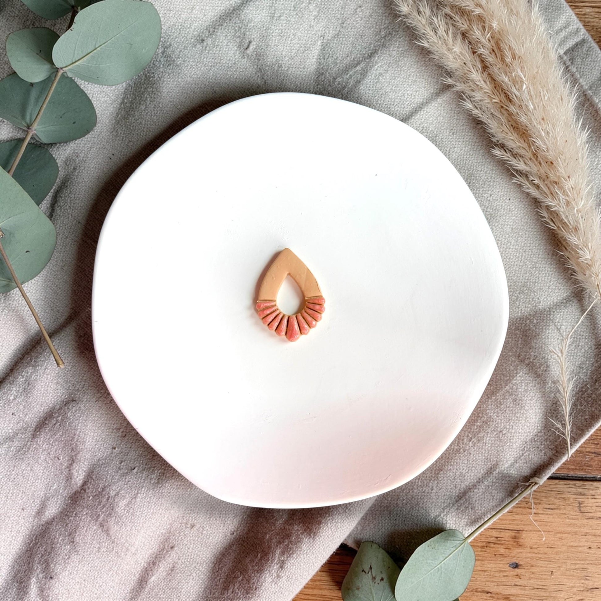 Small teardrop-shaped clay piece with fan texture at the bottom displayed on a white plate with neutral fabric and greenery.