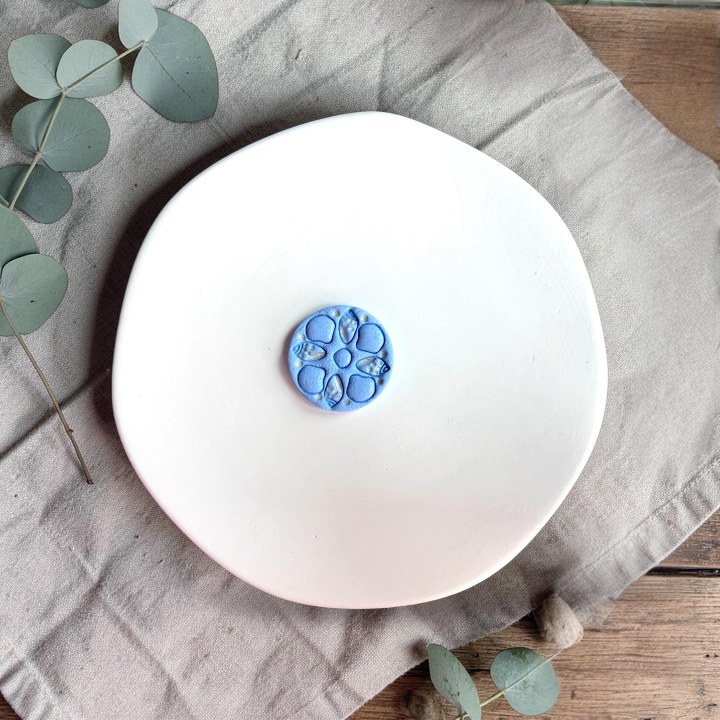 Blue finished clay piece with circular sea shell pattern placed on a white plate with neutral fabric and greenery.
