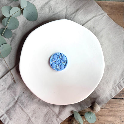 Blue finished clay piece with circular sea shell pattern placed on a white plate with neutral fabric and greenery.