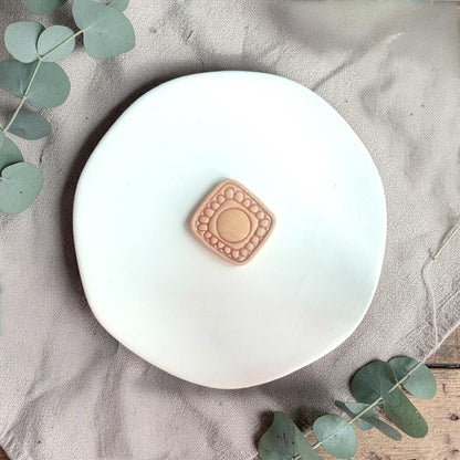 Square polymer clay cutter with raised circular center and decorative lace border placed on a white plate with neutral fabric and greenery.