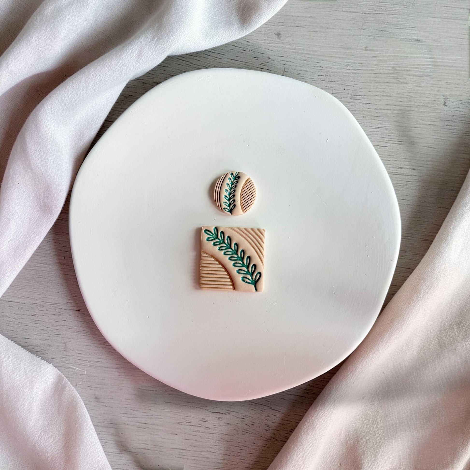 Finished clay pieces in square and circle shapes with textured leaf design placed on a white plate with neutral fabric.