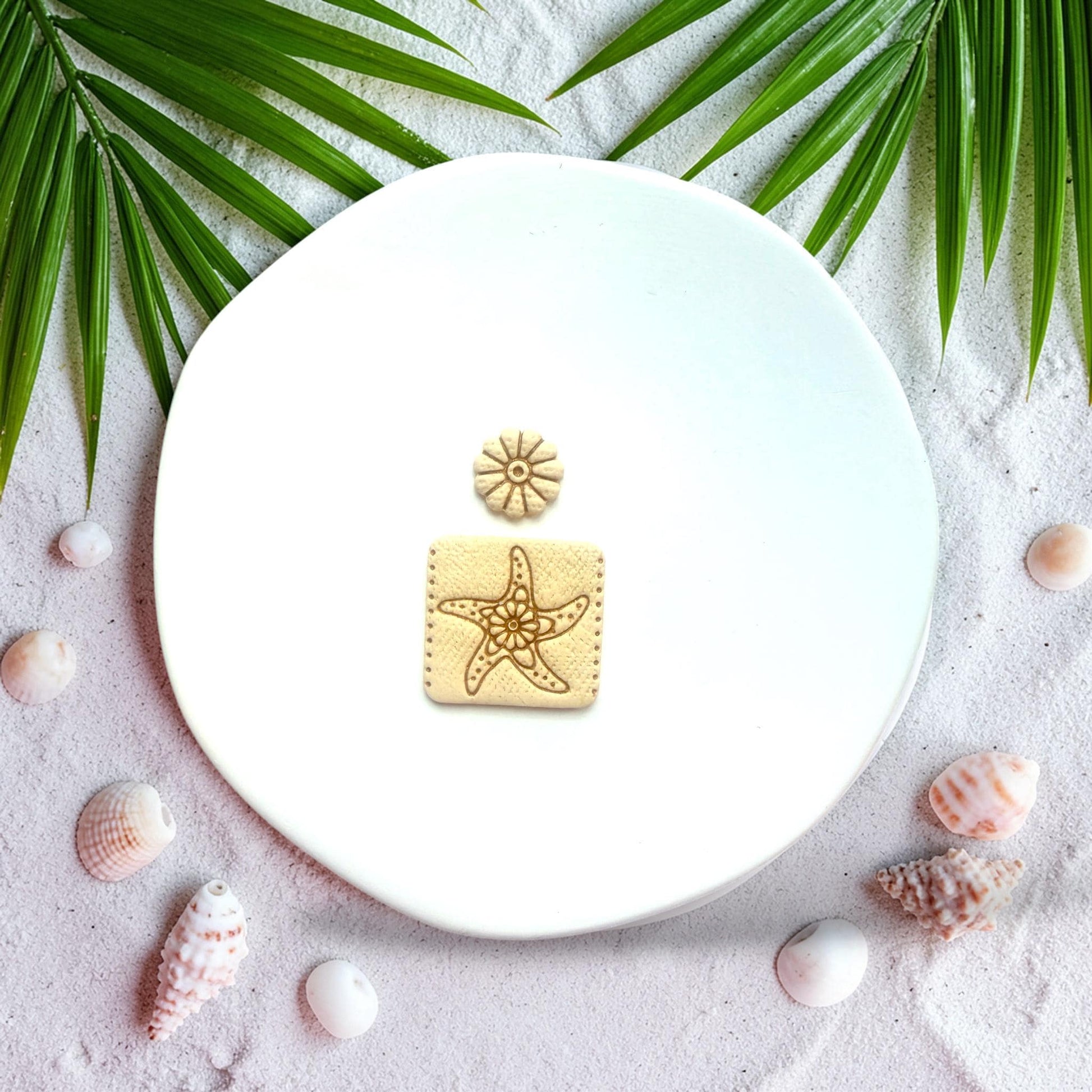 Starfish square polymer clay cutter with flower stud displayed on beach sand with shells and palm leaves