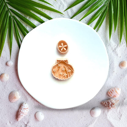 Floral scallop shell polymer clay cutter with daisy stud displayed on beach sand with seashells