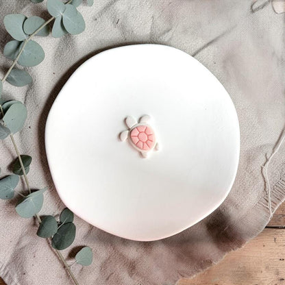 Sea turtle clay piece displayed on white plate with neutral fabric and greenery background