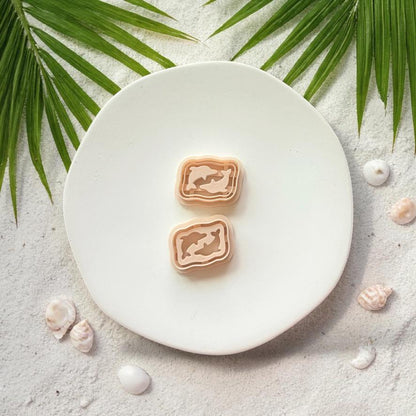 Dolphin rectangle clay cutters displayed on white plate with sand, shells, and palm leaves