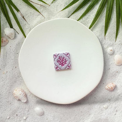 Purple square coral tile clay piece on white plate with sand, seashells, and palm leaves