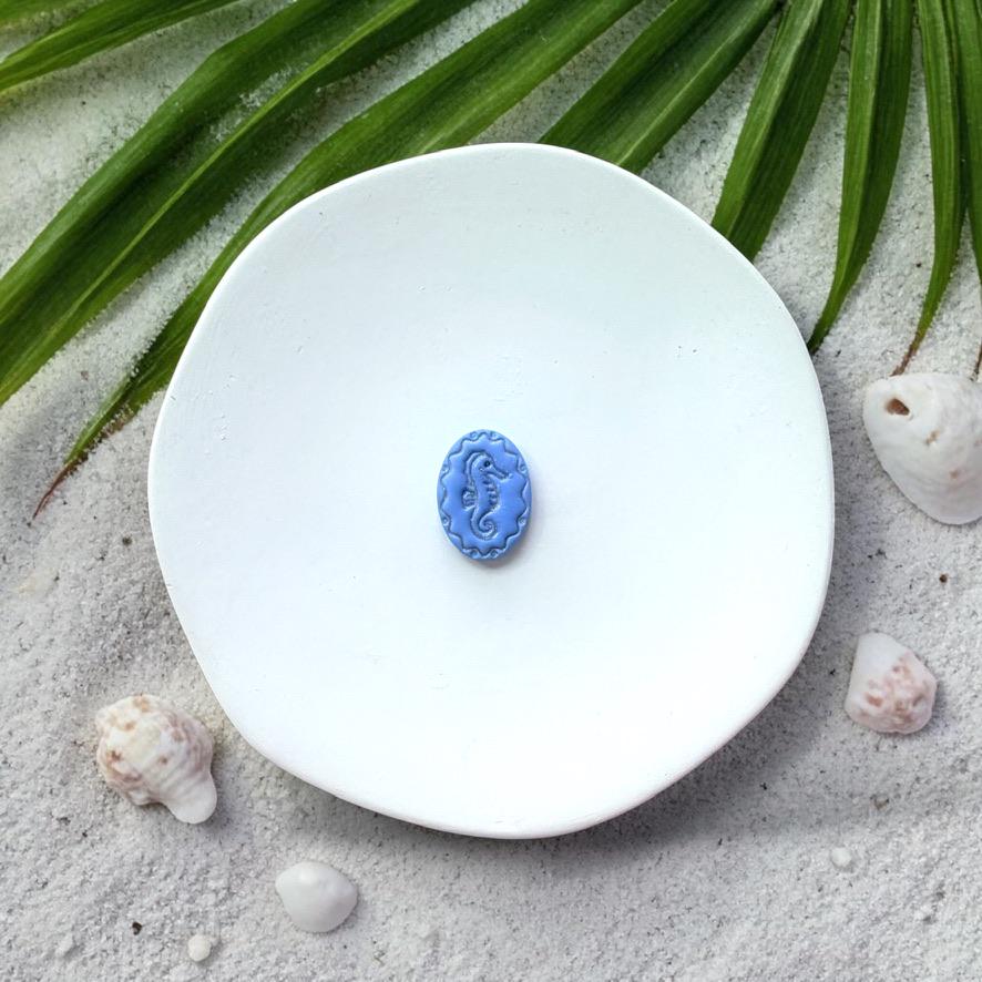 Blue oval seahorse clay piece on white plate with sand, seashells, and palm leaves