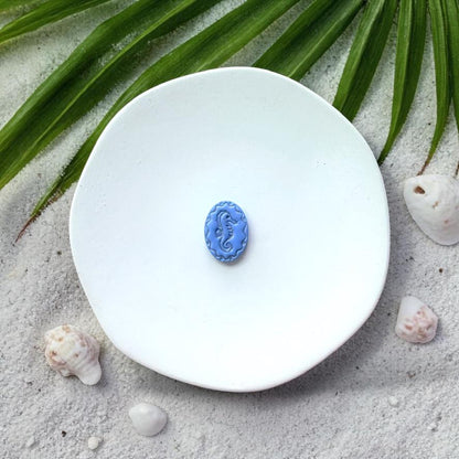 Blue oval seahorse clay piece on white plate with sand, seashells, and palm leaves