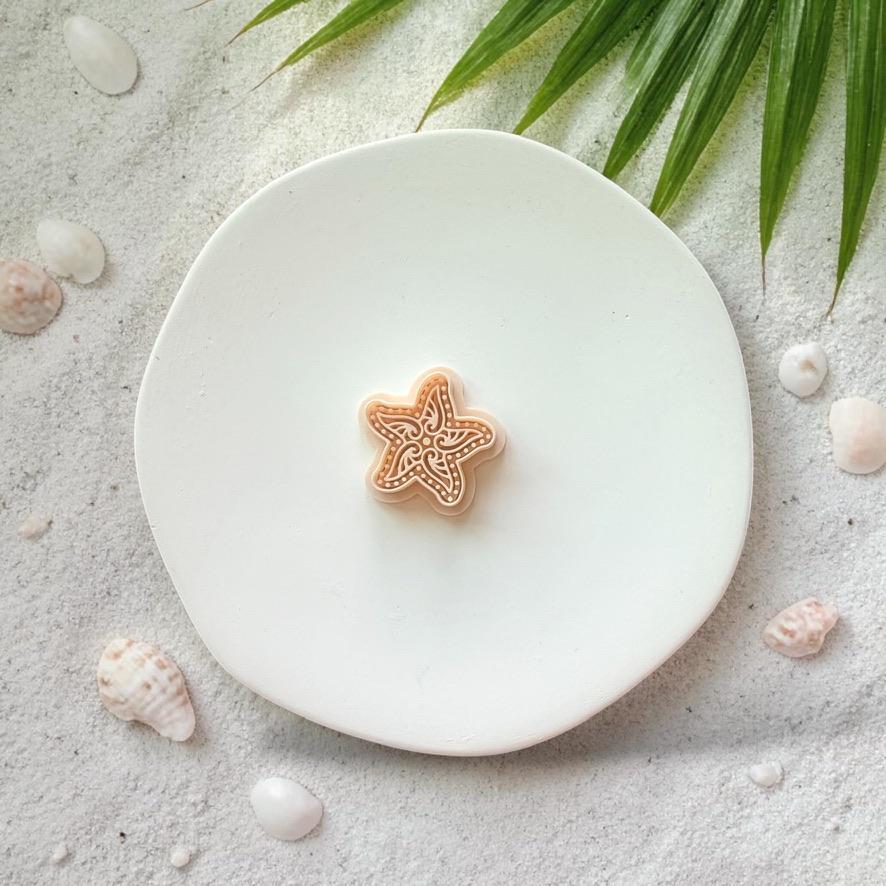Starfish clay cutter on white plate with sand, seashells, and tropical palm leaves