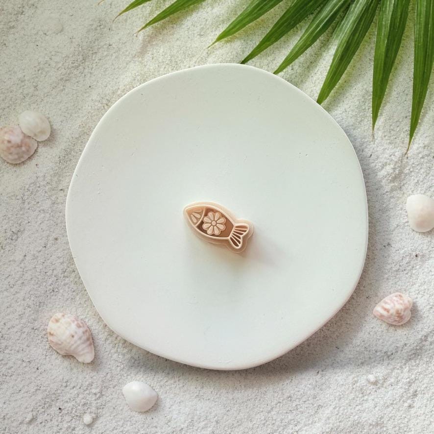 Fish clay cutter on white plate with sand, seashells, and tropical leaves