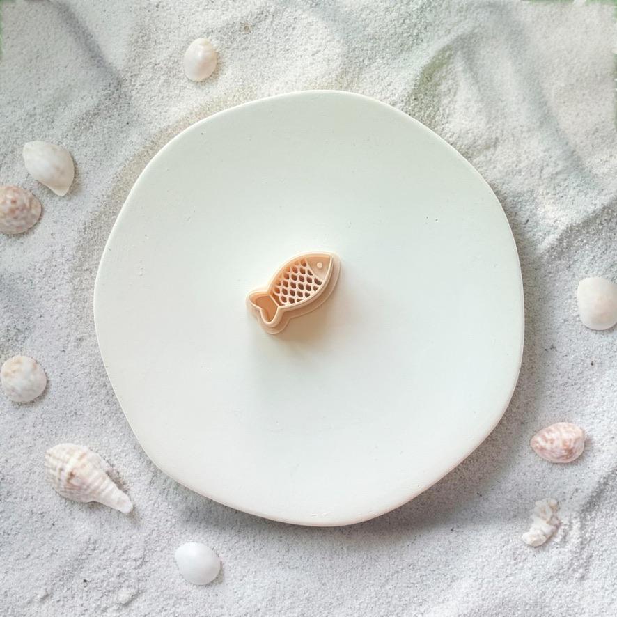 Fish clay cutter with scale detail on white plate with sand, seashells, and tropical leaves