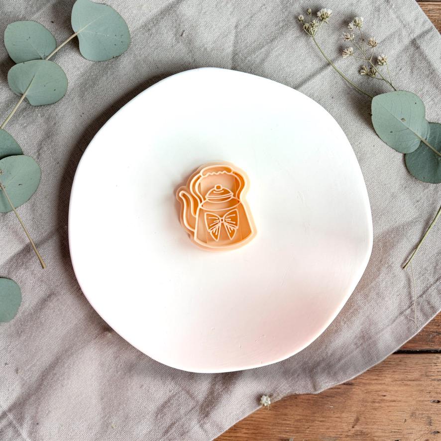 Teapot polymer clay cutter displayed on white plate with neutral fabric and greenery