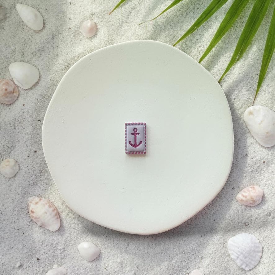 Small purple anchor tile clay piece on white plate with sand, seashells, and palm leaves