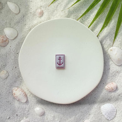 Small purple anchor tile clay piece on white plate with sand, seashells, and palm leaves