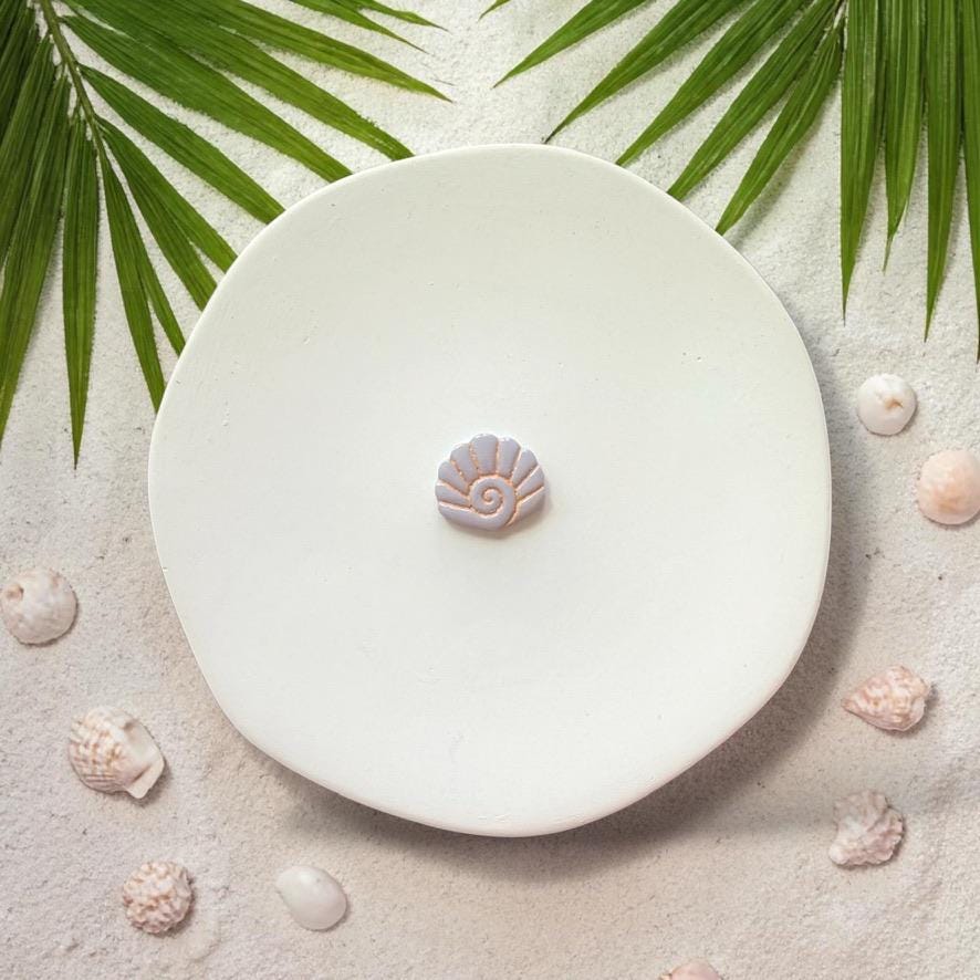 Small spiral seashell clay design on white plate with sand, shells, and palm leaves