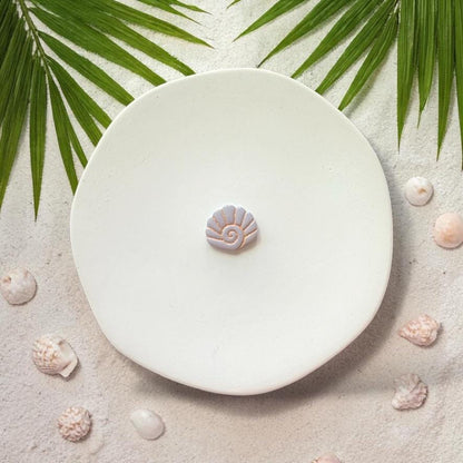 Small spiral seashell clay design on white plate with sand, shells, and palm leaves