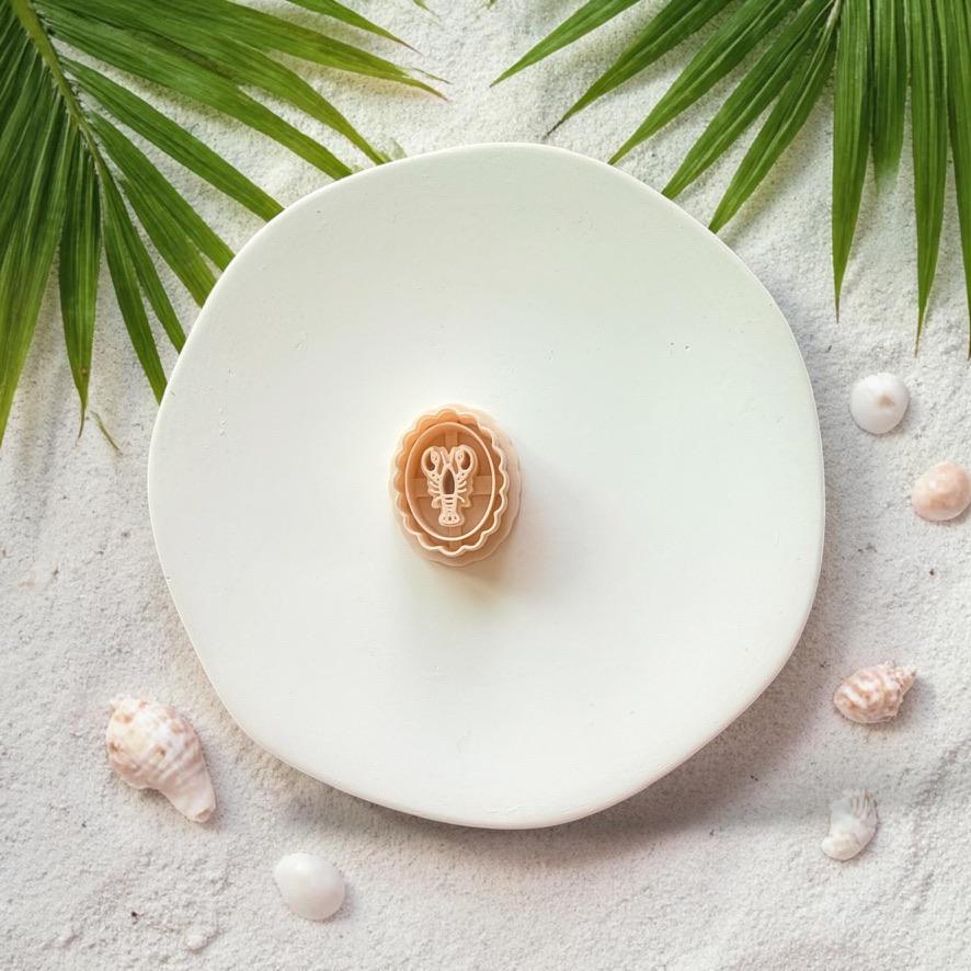 Lobster cameo clay cutter on white plate with sand, seashells, and tropical leaves