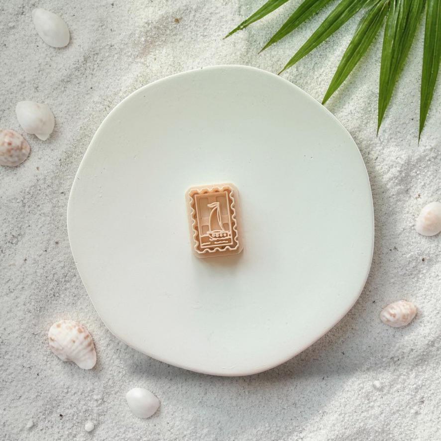 Sailboat frame clay cutter on white plate with sand, seashells, and tropical leaves