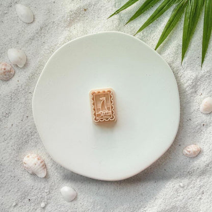Sailboat frame clay cutter on white plate with sand, seashells, and tropical leaves