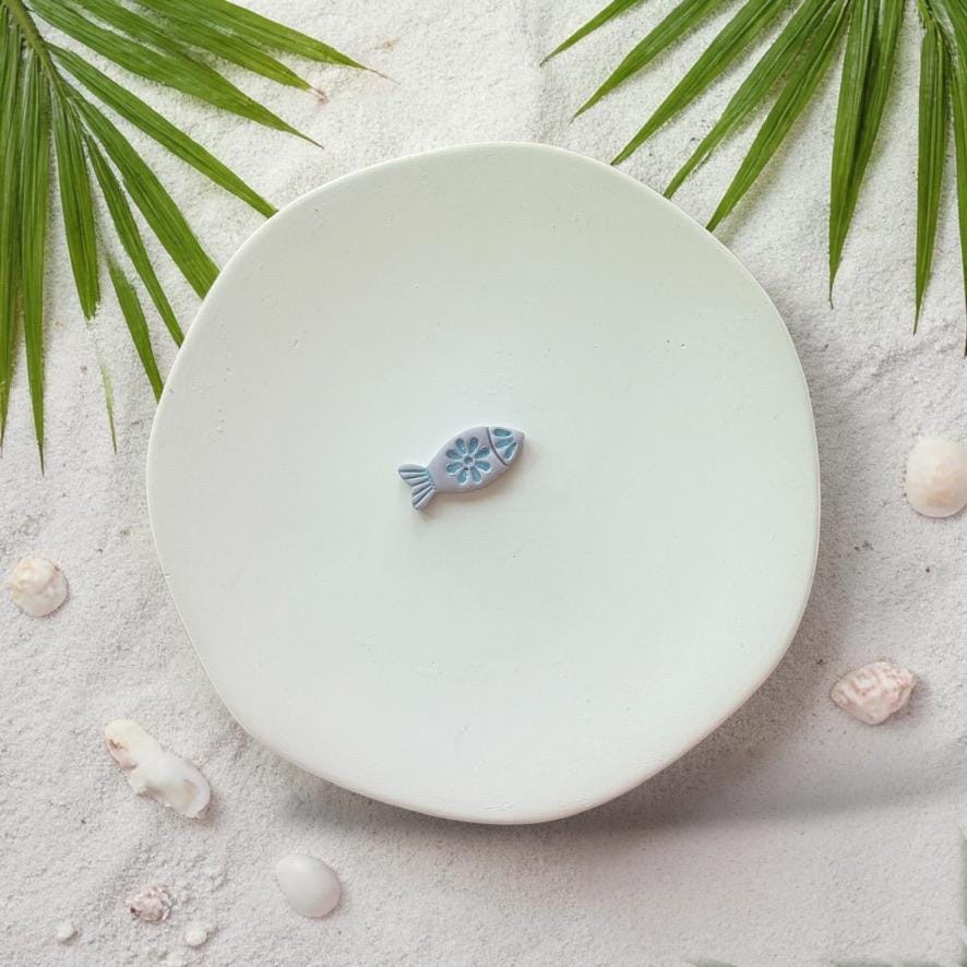 Finished fish clay piece in blue on white plate with beach sand and shells