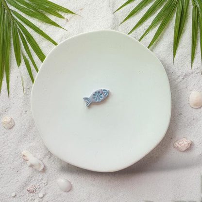 Finished fish clay piece in blue on white plate with beach sand and shells