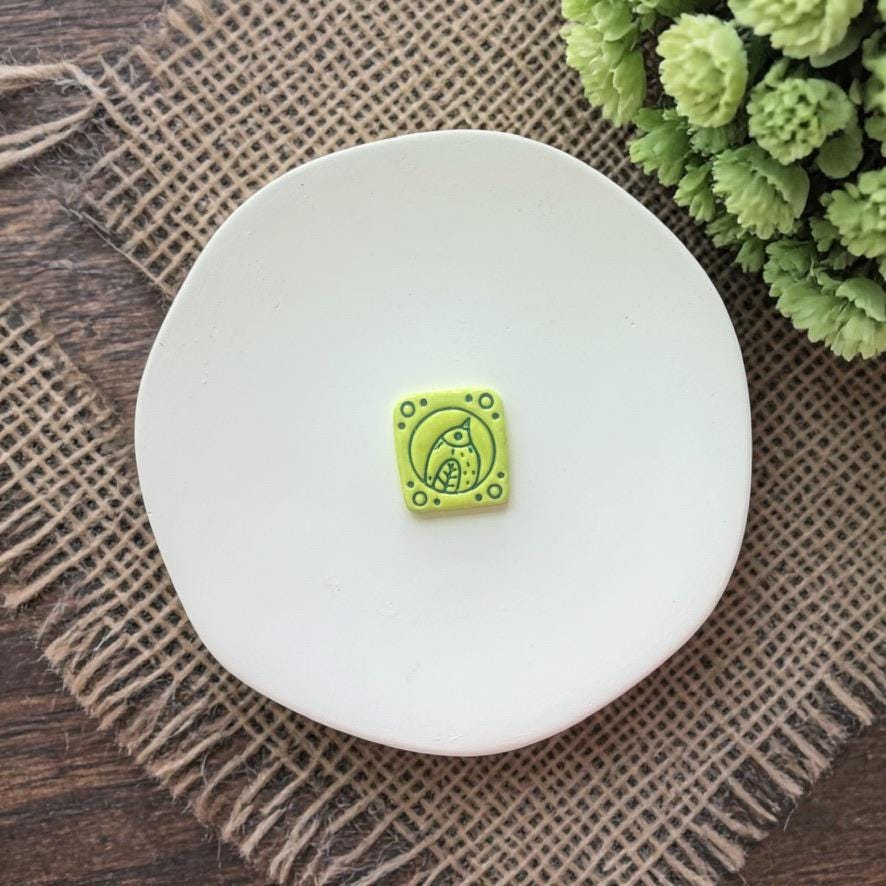 green clay bird tile design on white plate with rustic fabric background