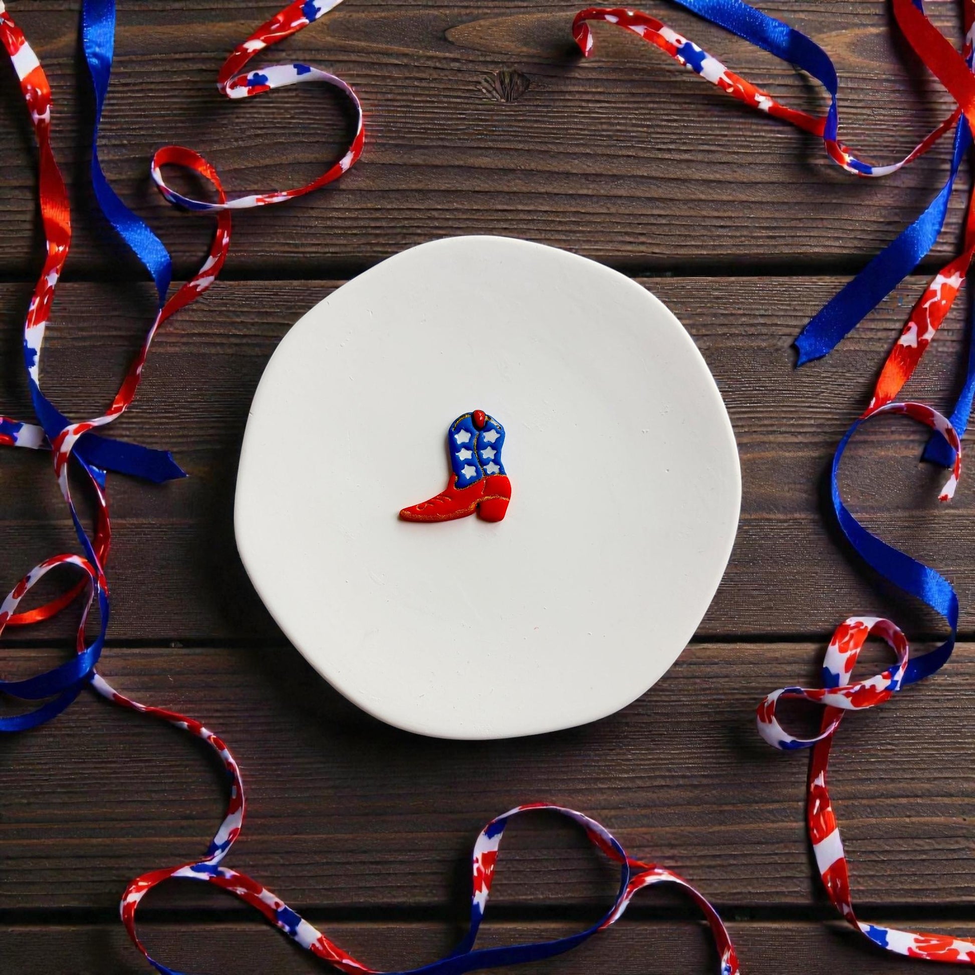 Finished red white and blue cowboy boot clay earring beside matching cutter on white display surface with patriotic ribbon accents