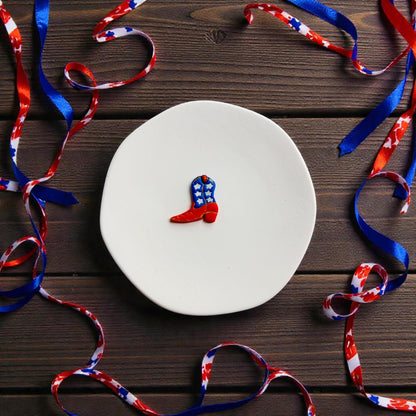 Finished red white and blue cowboy boot clay earring beside matching cutter on white display surface with patriotic ribbon accents