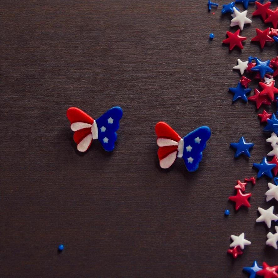 Pair of red white and blue patriotic butterfly clay earrings displayed on dark wooden background with star confetti