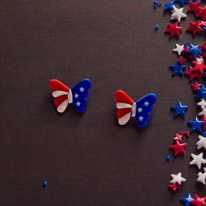 Pair of red white and blue patriotic butterfly clay earrings displayed on dark wooden background with star confetti