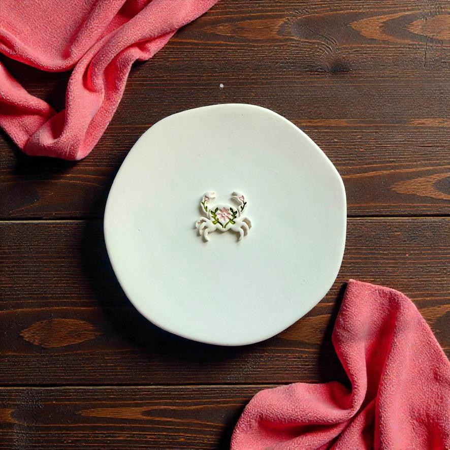 Finished folk inspired crab polymer clay earring displayed on white ceramic plate with pink coastal fabric background