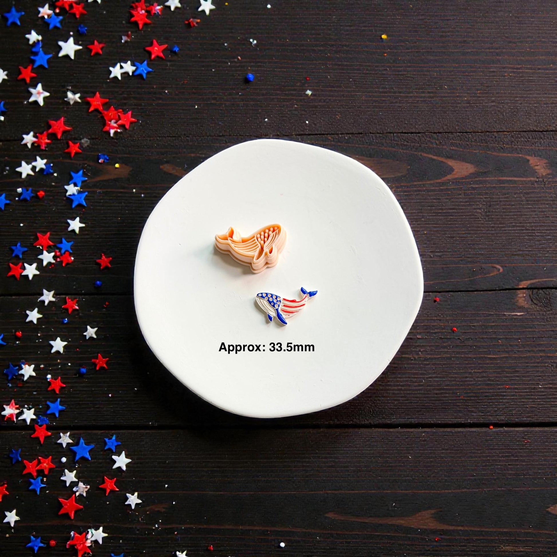 Patriotic whale polymer clay cutter and finished Americana whale clay piece displayed on white ceramic plate with red white and blue confetti