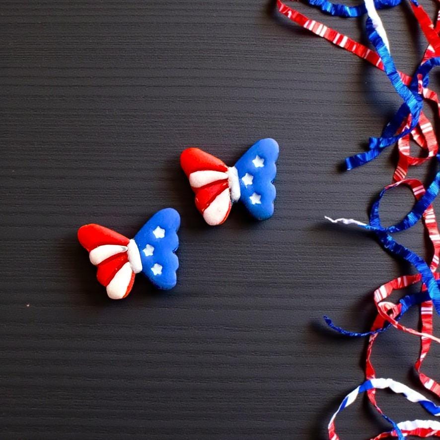 Pair of red white and blue butterfly clay earrings displayed on dark background with patriotic curling ribbon decorations