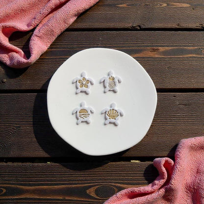 Finished tropical turtle clay earrings featuring flower, pineapple, sunset, and shell center designs displayed on white ceramic plate
