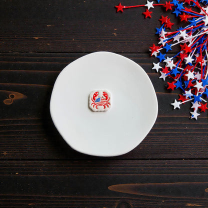 Finished patriotic crab clay earring in red white and blue colors displayed on white ceramic plate with patriotic star decorations