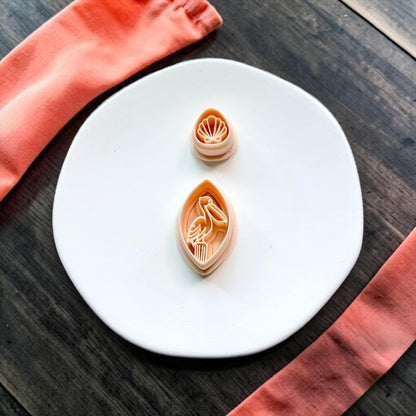 Pelican polymer clay cutter and matching seashell topper cutter displayed on a white ceramic plate with coral-colored fabric.