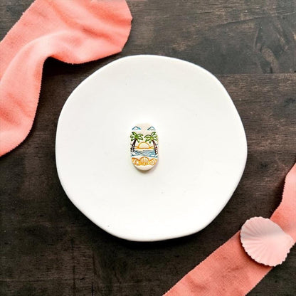 Finished tropical beach sunset clay piece displayed on a white ceramic plate with coral fabric and seashell accents.