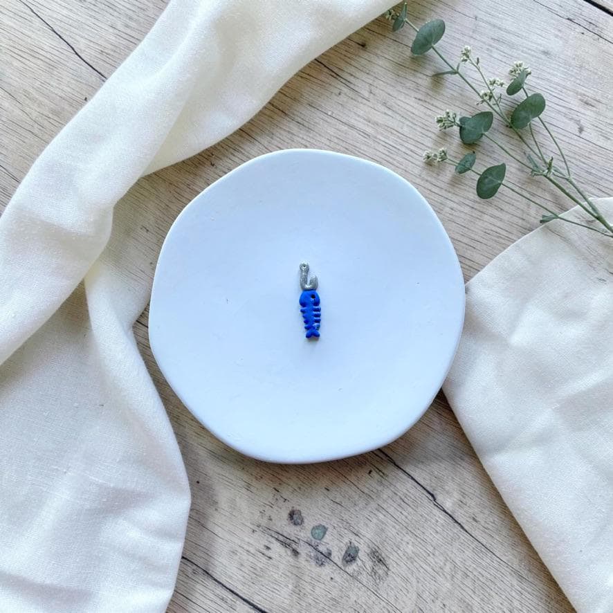 Finished blue fish bone and silver fishing hook clay charm displayed on a white ceramic plate with neutral fabric and greenery.