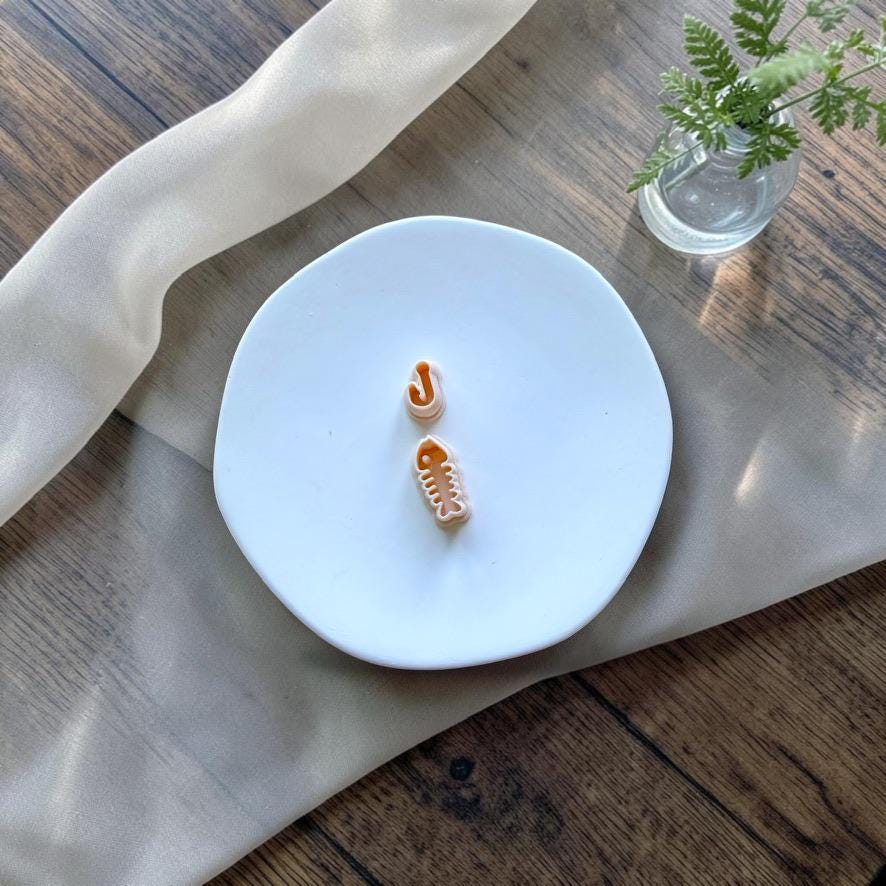 Fishing hook and fish bone polymer clay cutters displayed on a white ceramic plate.