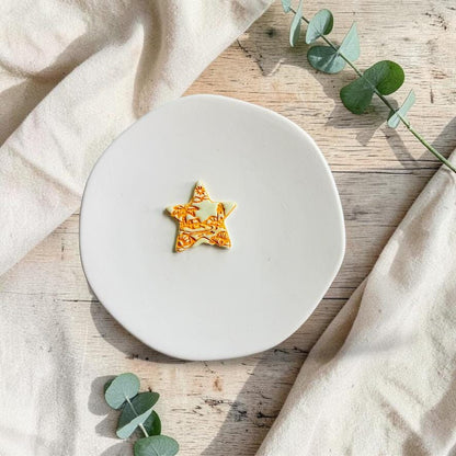 Finished star-shaped beach scene polymer clay earring on a white ceramic plate with eucalyptus accents.