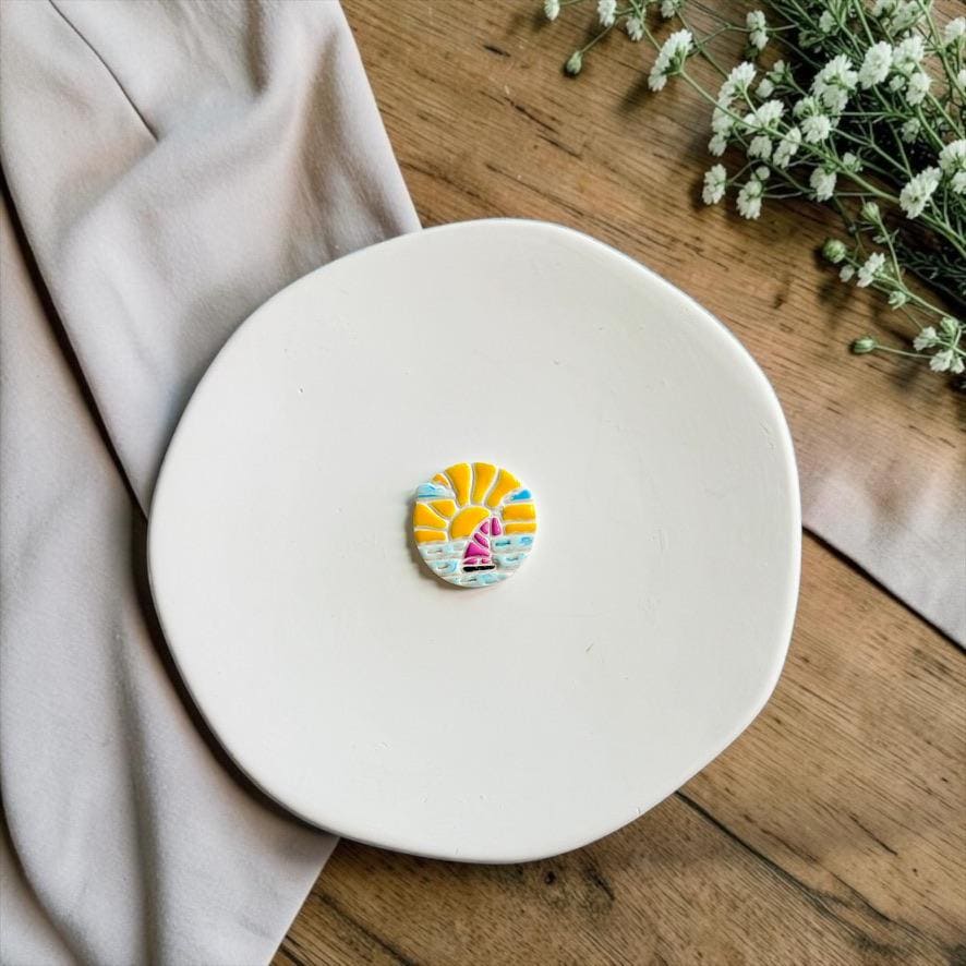Sunset sailboat polymer clay cutter displayed on a white ceramic plate with neutral fabric and white flowers.