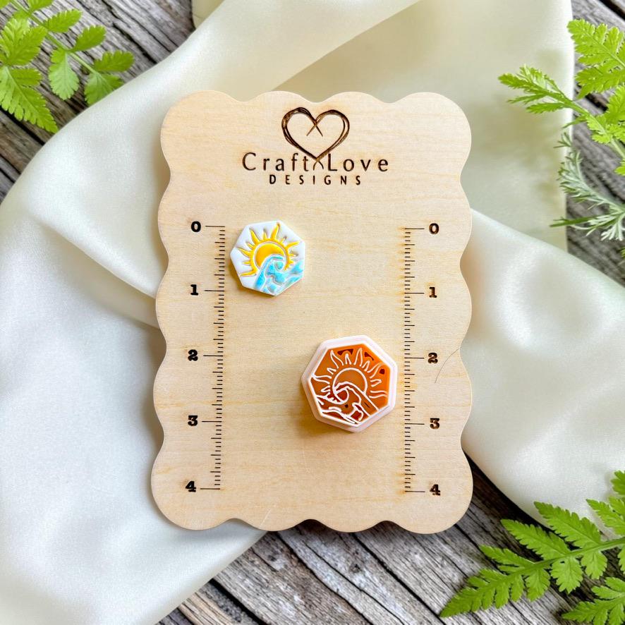 Finished hexagon polymer clay earring featuring a sun rising over ocean waves displayed beside the matching cutter on a Craft Love Designs sizing board.
