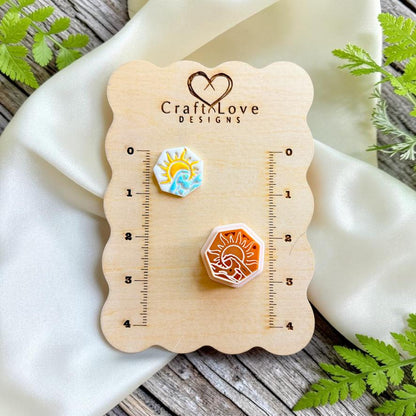 Finished hexagon polymer clay earring featuring a sun rising over ocean waves displayed beside the matching cutter on a Craft Love Designs sizing board.